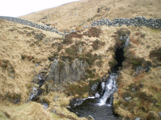 irregular land covered with golden, dried grass, crossed by a dry-stone wall and with a stream bubbling out of a dark crack in the ground
