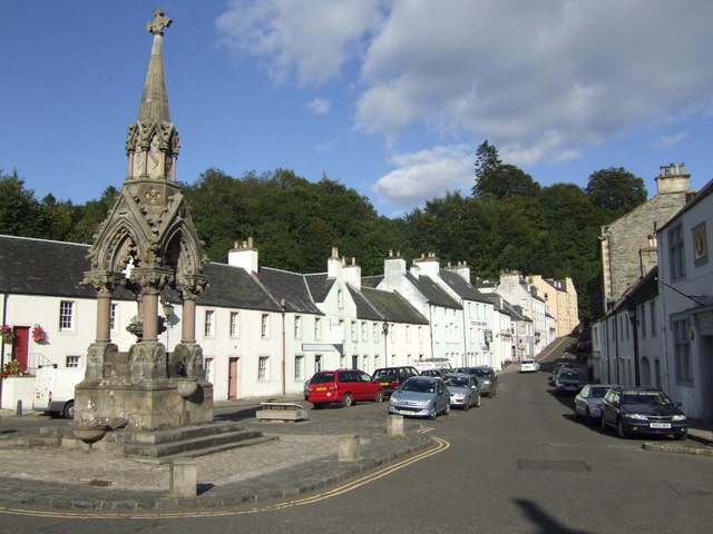 a village high-street rising up a slope into the distance, with a small, elaborately-carved stone spire supported on four pillars in the foreground, and an assortment of small squarish white buildings along either side of the street