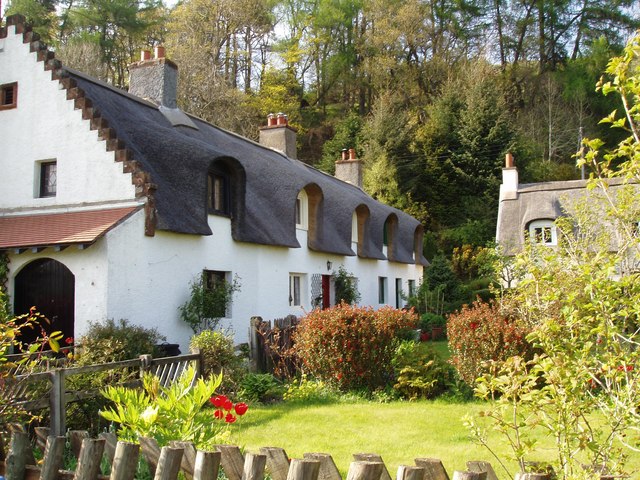 terraced row of two-storey white buildings with dormer windows surrounded by smooth, elaborate dark thatch, with a bright garden in the foreground and another thatched building in the background
