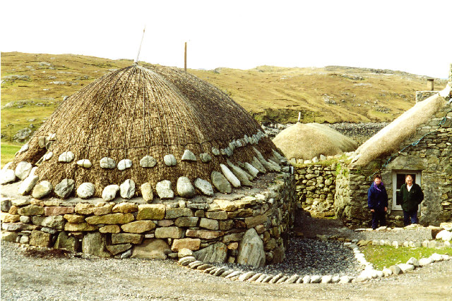 low, rough stone building with a thatched roof which doesn\'t overhang the thick walls but stops before reaching the edge of them, the thatch being weighted down by a net with large stones hanging from the edge all the way round