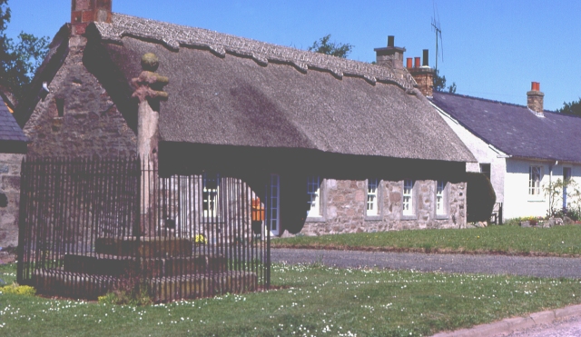 long low grey stone building, possibly a Post Office, with a thatched roof with fancy trim along the roof-ridge: in front is a stretch of grass and a fenced monument consisting of steps up to a short pillar topped by two horizontal sheets with curled edges, and a ball finial