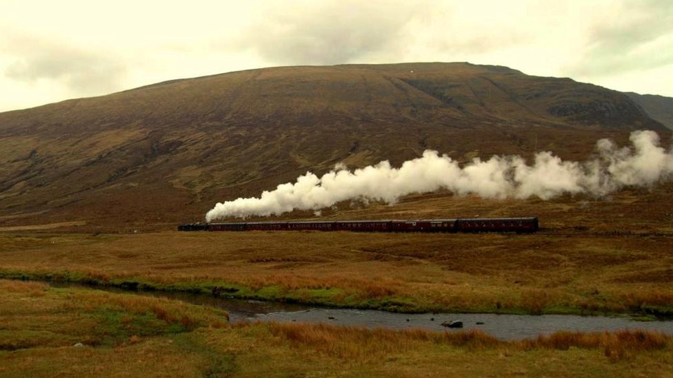 long, panoramic view of a steam-train steaming across a bleak valley carpeted with bracken, with a bare hill behind it and a stream in the foreground