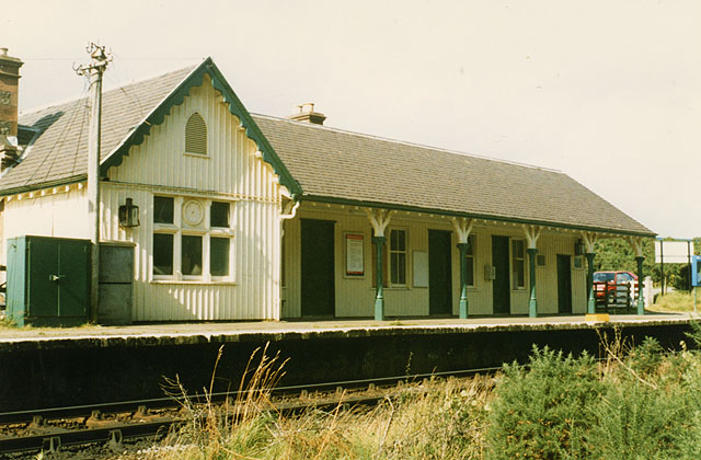 station building painted cream with green trim, with vertical ribs indicating that the walls are either corrugated iron or weatherboard, having a gabled, house-like section to the left and then a long portion stretching along the platform to the right, this section having a roof which extends forwards and acts as a partial canopy to the platform