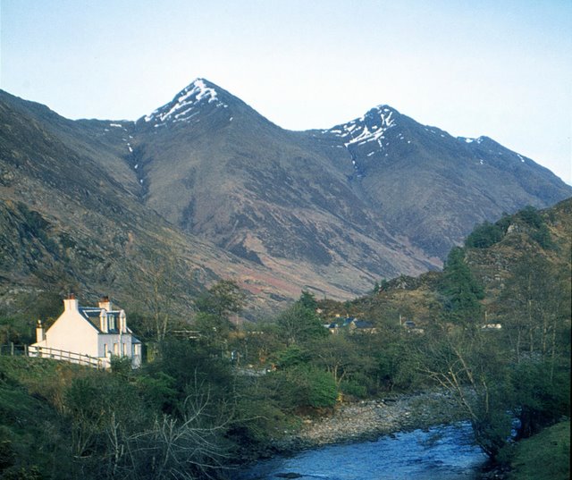 a small white house and part of a river in the foreground, more buildings glimpsed through trees in the middle ground and two jagged mountain peaks in the background