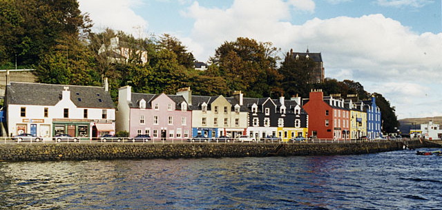 view across the open water of a harbour towards a quayside and a row of squarish houses and shops painted in bright colours