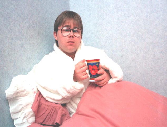 Photo' of Eric Cullen sitting up in bed, in white towelling dressing-gown, holding cup of tea