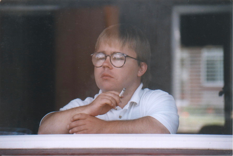 Eric Cullen in three-quarter view seen through window, holding cigarette up near face