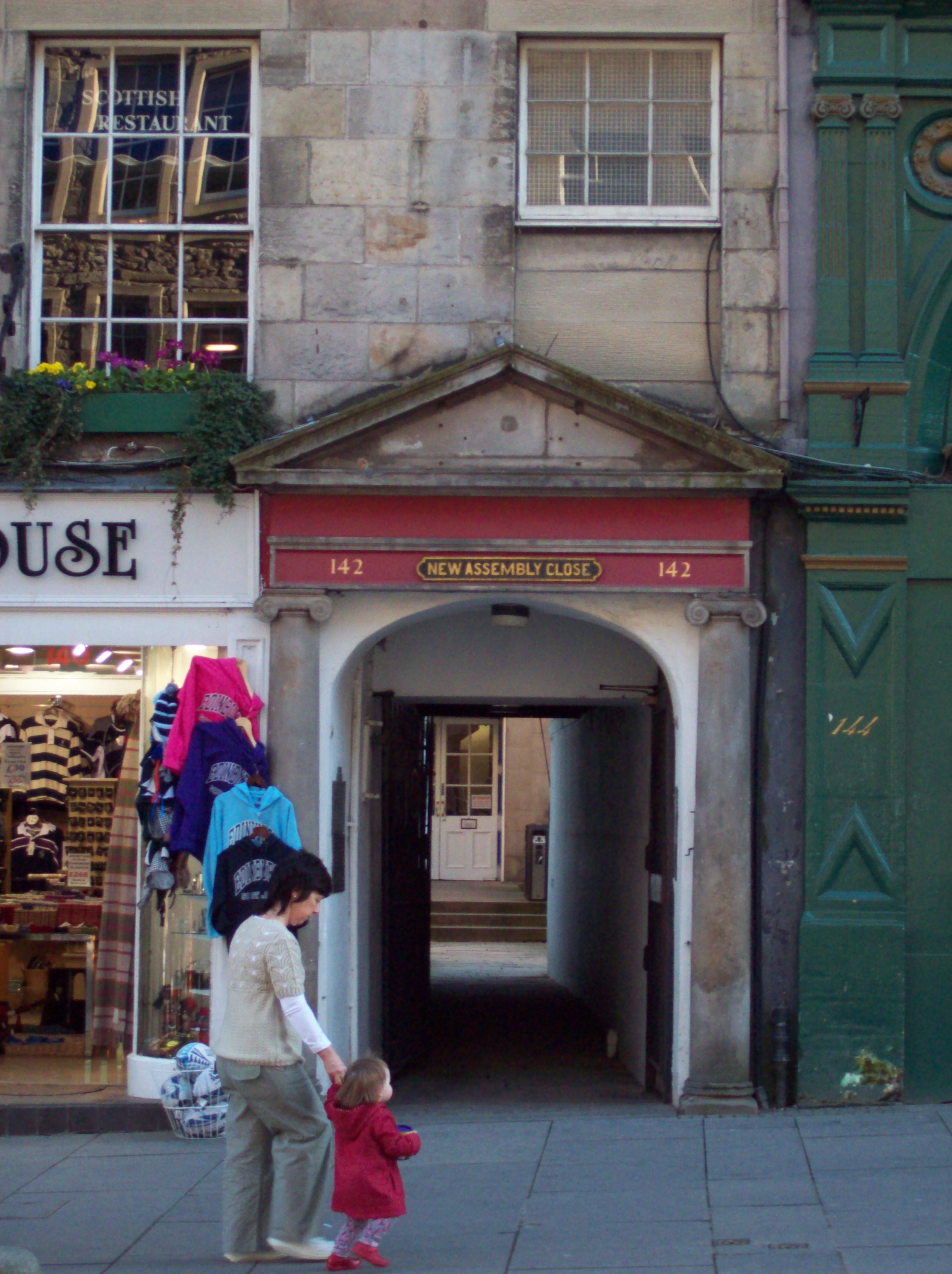a white archway flanked by grey stone classical pillars, opening between shops and into a tunnel through the building, with a red strip above it bearing the label \'NEW ASSEMBLY CLOSE\' and a triangular stone architrave above that: an open courtyard and a white dorway with steps up to it can be glimped through the tunnel