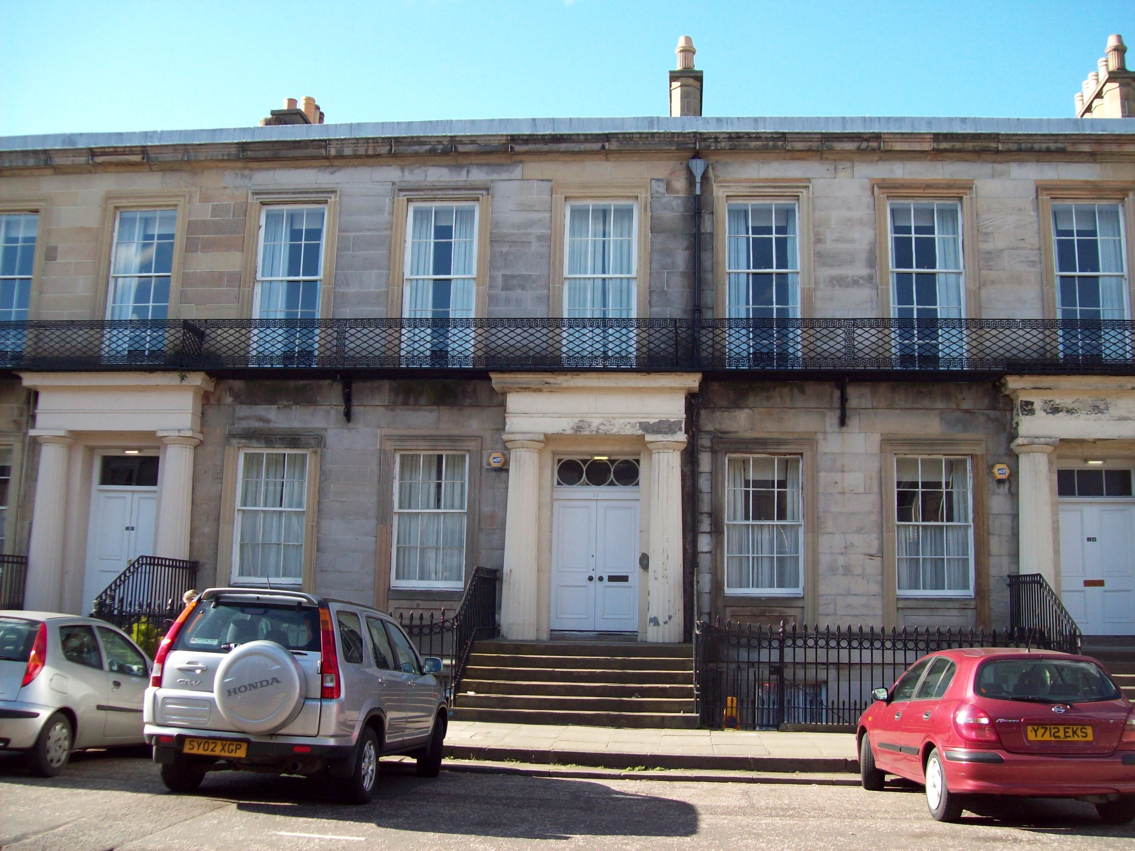 row of three-storey sandstone buildings having a basement storey partially below ground but visible through iron railings; then a ground floor with large windows and with double doors framed by classical porticoes painted cream, with flights of steps leading up from a pavement which slopes down to the right; then an upper floor with very large floor-to-ceiling windows, in front of which is a low black-painted metal grille running the length of the street and probably intended to hold window-boxes