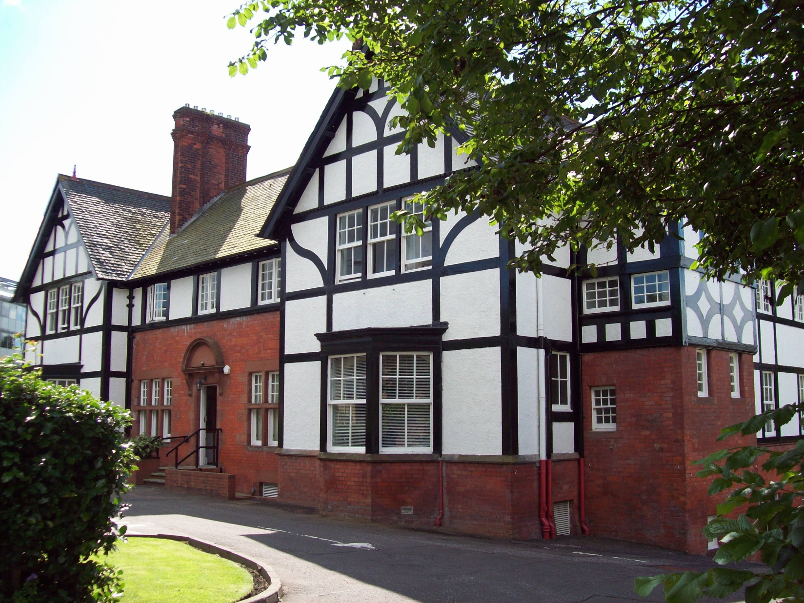 large mock-Tudor building having an end-on, black and white gable-end section to ether side and in the middle a red brick stretch with high chimneys and a band of black and white along the upper floor, all set in extensive grounds with a high hedge in front