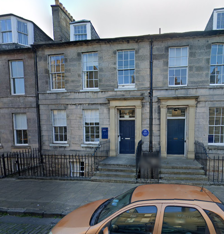 blocky grey stone building with white windows divided into multiple small panes, with a below-ground \