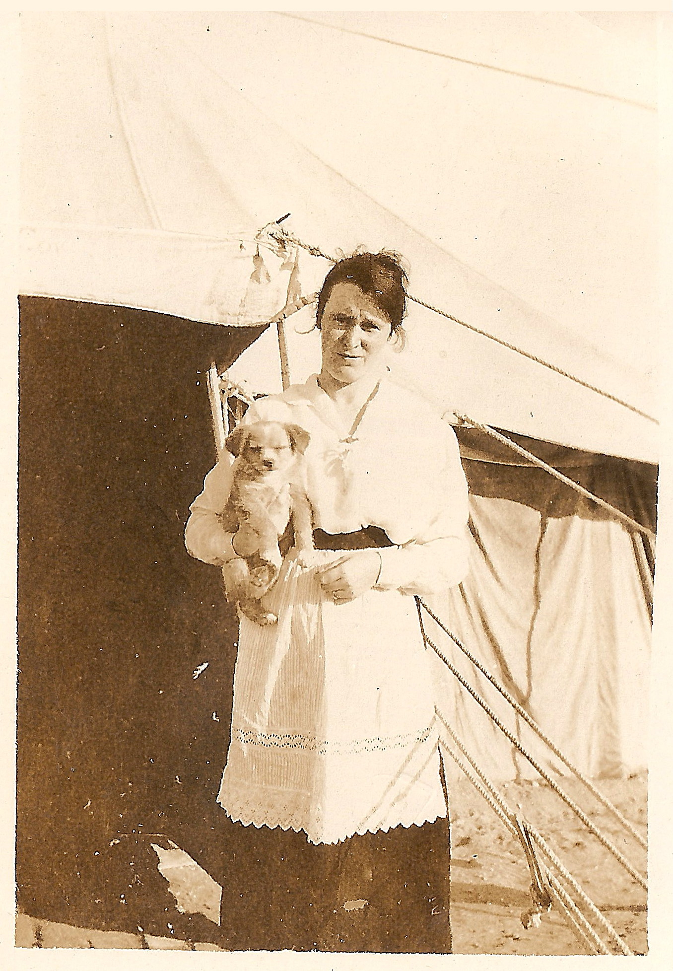 dark-haired woman in a long dark skirt and a white smock or apron, holding a puppy and standing in front of a tent