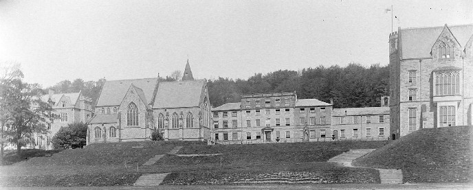 black and white photo\' of a long skein of tall, old-fashioned stone and brick buildings at the top of a stepped grassy slope, with a church at the centre and a gentle rise of hillside behind them