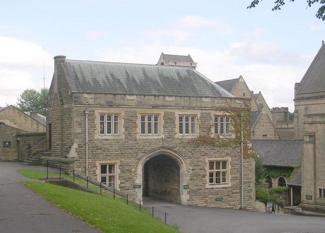 two-storey building in grey-brown brick, with Mediaeval-style windows and a huge arched tunnel leading through the centre of the building: beyond it to the right is a cluster of abbey buildings and in the left foreground a path at a higher level cuts across the picture