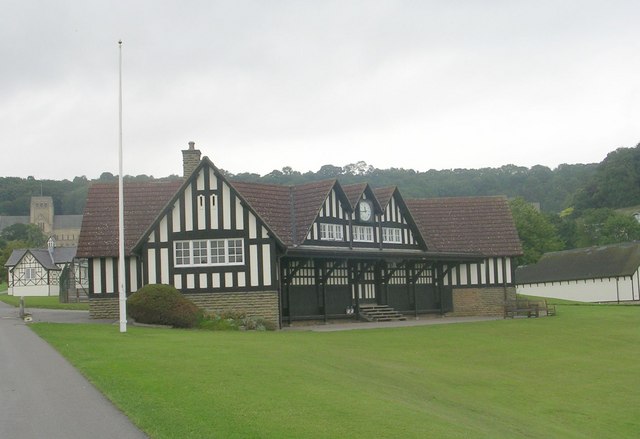 medium-sized black, white and brown mock-Tudor building in a complex shape, set in  a playing field, with a recessed front under an upper floor supported on beams, and with wooden steps up to the door: behind it is a smaller mock-Tudor building in grey and white, and then the Abbey church with a wooded slope behind it