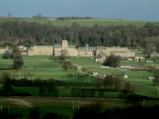 view across and down a wide expanse of green playing fields towards a long sprawl of large beige buildings, including a church with a square tower at centre: beyond them a wood climbs halfway up the slope of a high rolling green hill