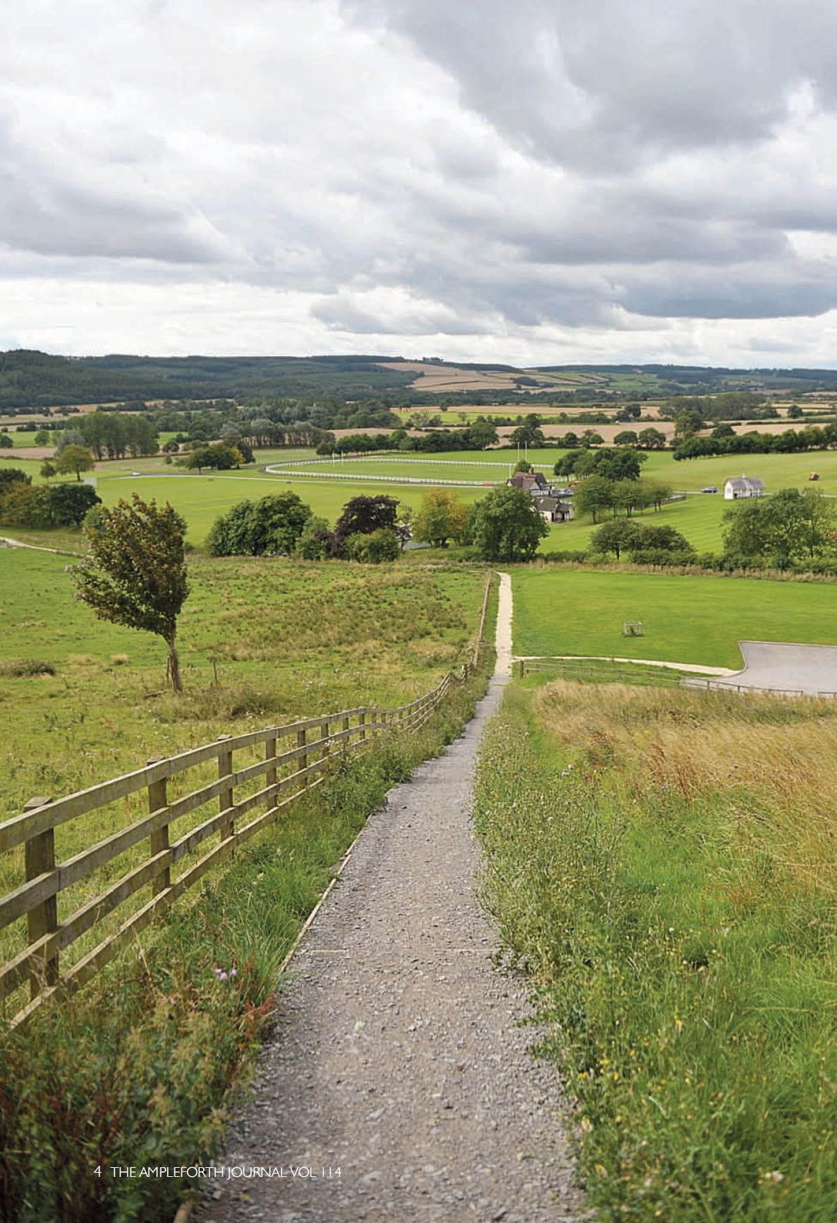 long view down a gravel path with a wooden fence at the left side, down a grassy slope and across a wide expanse of playing fields to low, rolling wooded hills in the distance