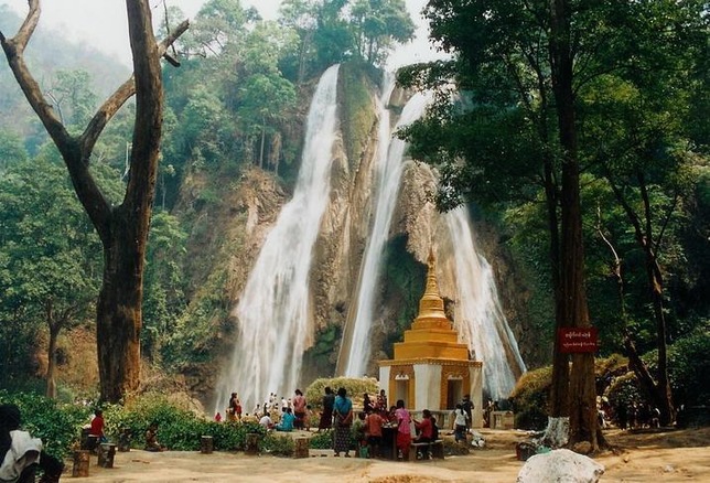 colour photo\' showing three skeins of water pouring down a rock, at the foot of which are tall trees, a small gold and white pagoda and a small crowd of visitors in bright clothes