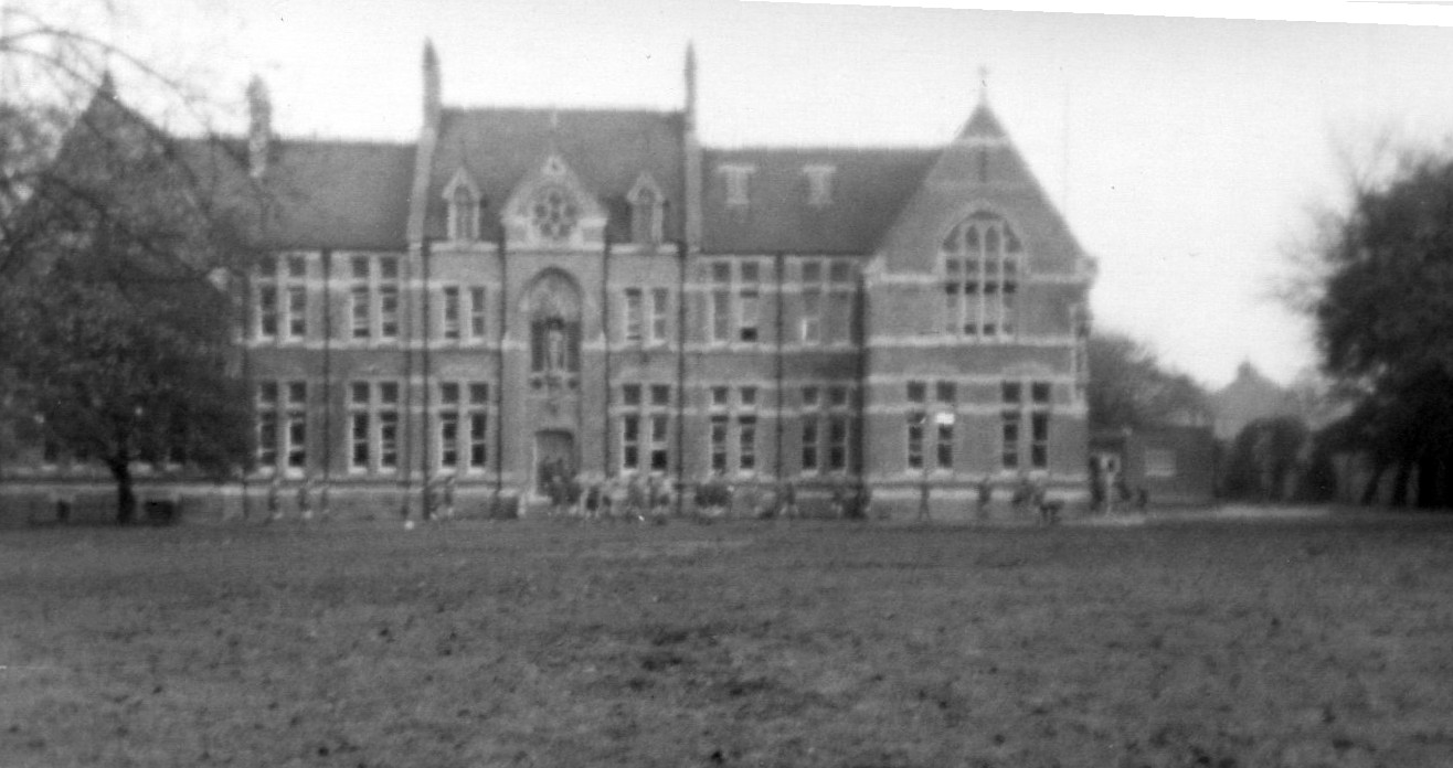 greyscale photo\' of a large Victorian Gothic building banded in red and cream brick, with tall windows and a complex roof