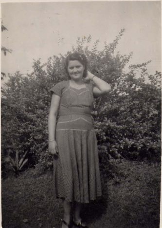 black and white photo\' of a slightly plump, dark-haired woman aged about thirty-five, standing in a garden with her left hand up behind her head, and wearing a loose, mid-calf-length summer dress