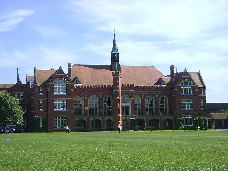view across a lawn to a large three-storey Victorian Gothic building in red brick with spires and turrets, with large cream-framed windows, square on the end wings and arched along the central block, broken into lots of little panes