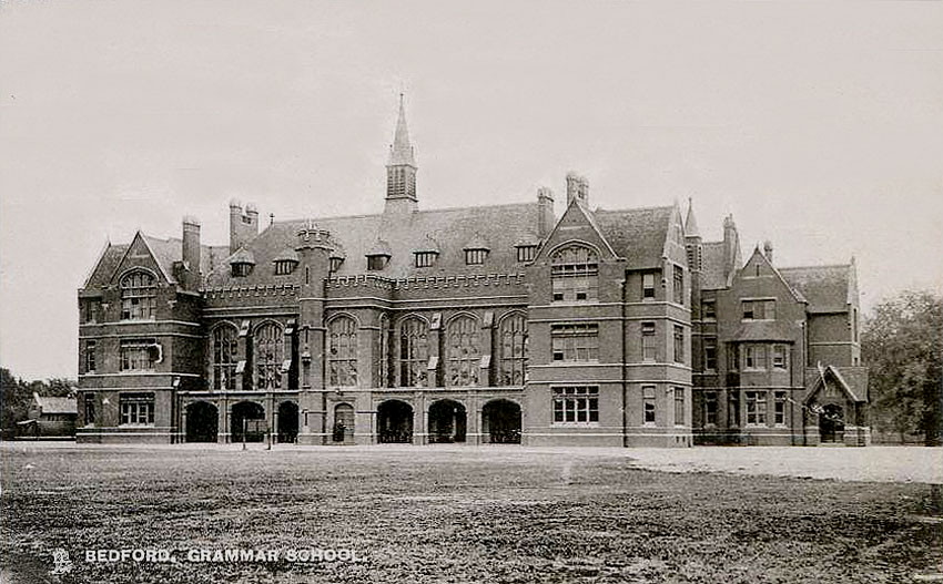 greyscale view across a lawn to a large three-storey Victorian Gothic building with spires and turrets, with large windows, square on the end wings and arched along the central block, broken into lots of little panes