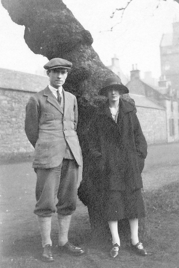 greyscale photo\' of a tall young man in plus-fours and tweeds and a tweed cap, and a young woman in a long dark coat, a wide-brimmed hat and shoes with fancy buckles, leaning against a tree with a long blank barn-like building behind it