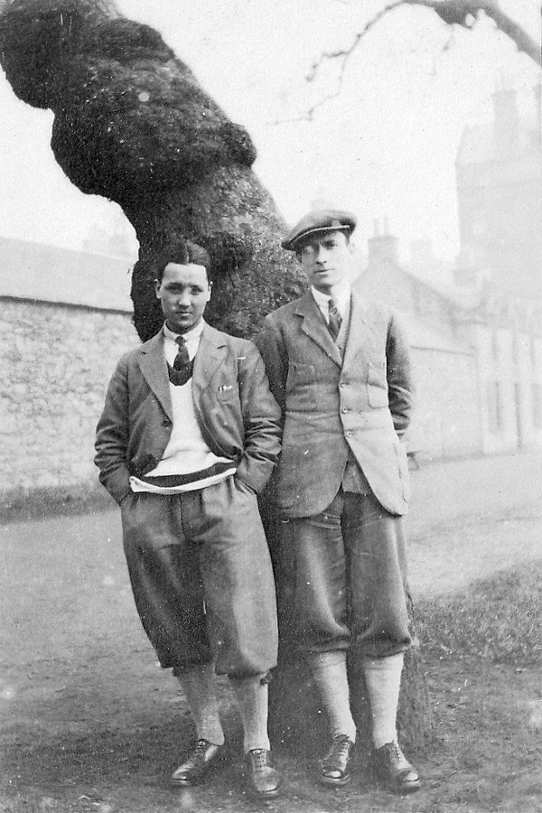 greyscale photo\' of two young men in plus-fours and tweeds, leaning against a tree with a long blank barn-like building behind it: the one on the left is short with centre-parted dark hair; the one on the right is tall and wearing a tweed cap