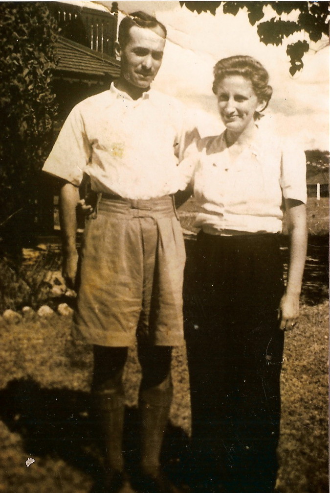 sepia photo\' of a slightly-built, dark-haired man wearing a loose white short-sleeved shirt, khaki shorts and hairy socks and a slim woman with a white short-sleeved shirt, dark trousers and Marcel-waved brown hair, standing with their arms round each other\'s waists in front of an outdoor scene of buildings and trees