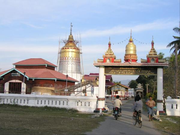 view past bicyclers along a concrete path leading under a square white gate with three mini golden pagodas on top and a brown and gold sign above the roadway: a white stone fence extends to either side of the gate and behind it on the left is a building with a two-tier red-tiled roof, and beying that a silver and gold tower resembling an oast-house and surrounded by scaffolding