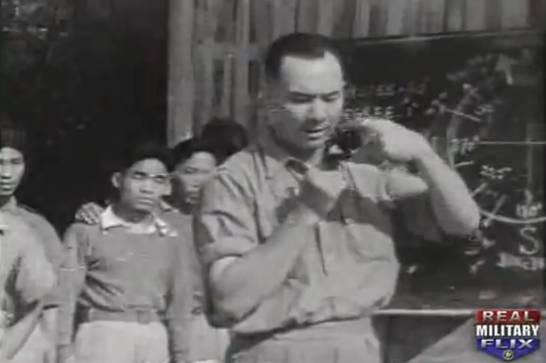 greyscale photo\' showing the upper body of a strongly-built man in an army shirt, with dark receding hair, with behind him a group of Asian soldiers wearing pullovers
