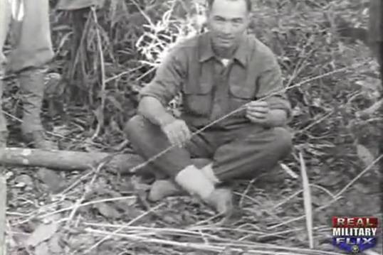 greyscale photo\' showing a strongly-built man in an army shirt, with dark receding hair, sitting cross-leggged on the jungle floor and holding a thin strip of bamboo