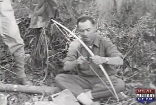 greyscale photo\' showing a strongly-built man in an army shirt, with dark receding hair, sitting cross-leggged on the jungle floor and holding a bow