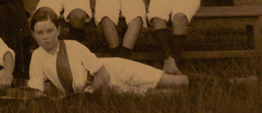 sepia photo\' showing a boy wearing white shirt and shorts with a diagonal, coloured sash across his chest, sprawled on the grass