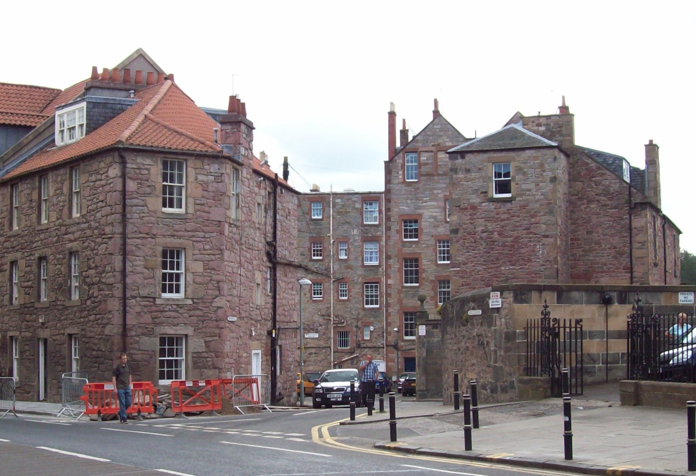 looking down a lane between a three-storey brown stone bulding on the left and a stone wall on the right: in the distance the lane splits, with the left fork leading into an enclosed yard with five-storey buildings of pale brown stone with red-brown trim round the windows, and the right fork winding away out of sight
