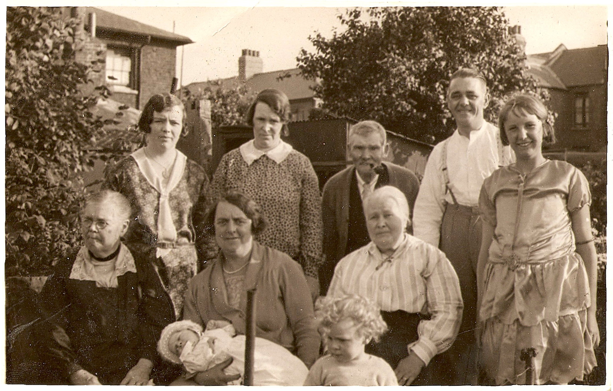 black and white photo\' of mixed group of men, women and children in 1920s clothing in a garden, ranging from an infant to very old, with in the front row three seated women visible from the waist up plus a standing girl to the right, with two men and two women standing behind them: the middle one of the seated women is holding a baby and there is a toddler with a mass of blond curls standing in front of all
