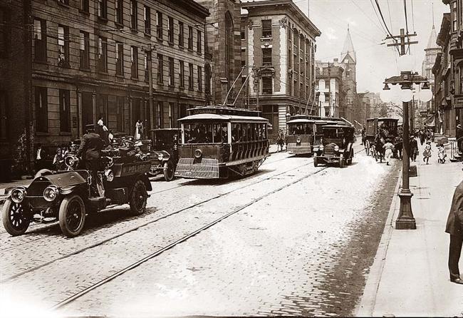 greyscale photo\' of a street-scene of trams and early motor-cars on a wide road lined with grand Victorian shop fronts