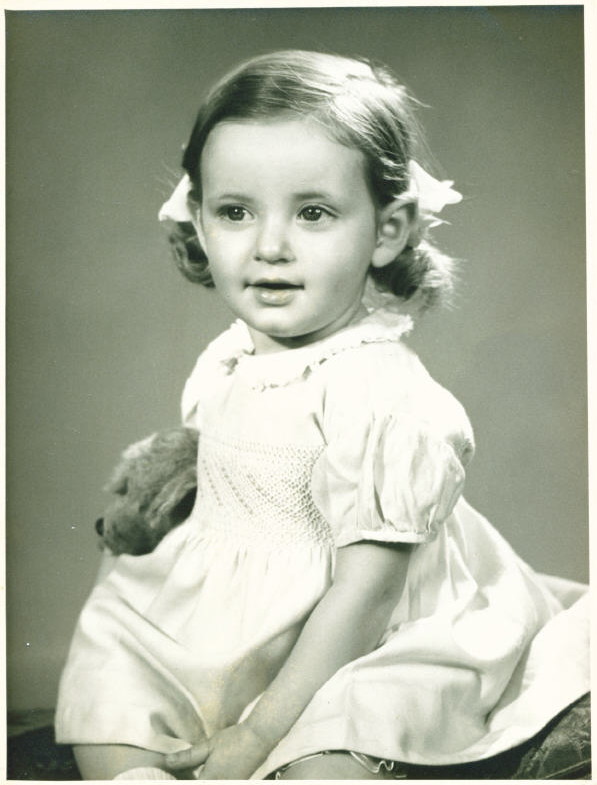 round-faced little girl about eighteen months old with light-brown hair tied in symmetrical bunches with bows on, wearing a loose white frock and sitting holding a grey toy poodle