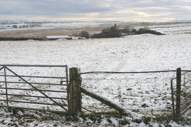 view from a metal gate across snowy fields towards farm buildings tucked down into a dip in the land