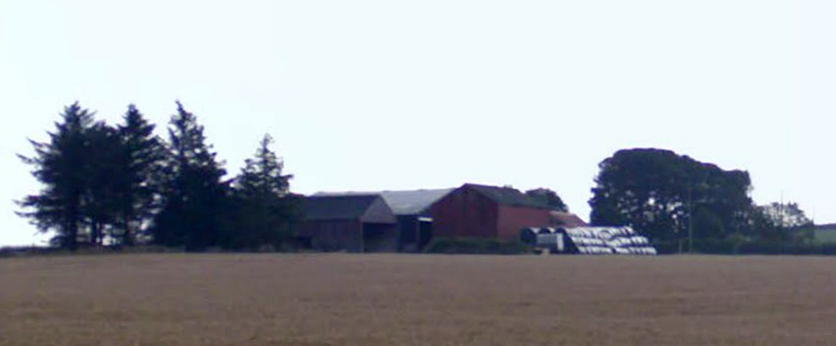 View across a brown field to a cluster of farm buildings among trees