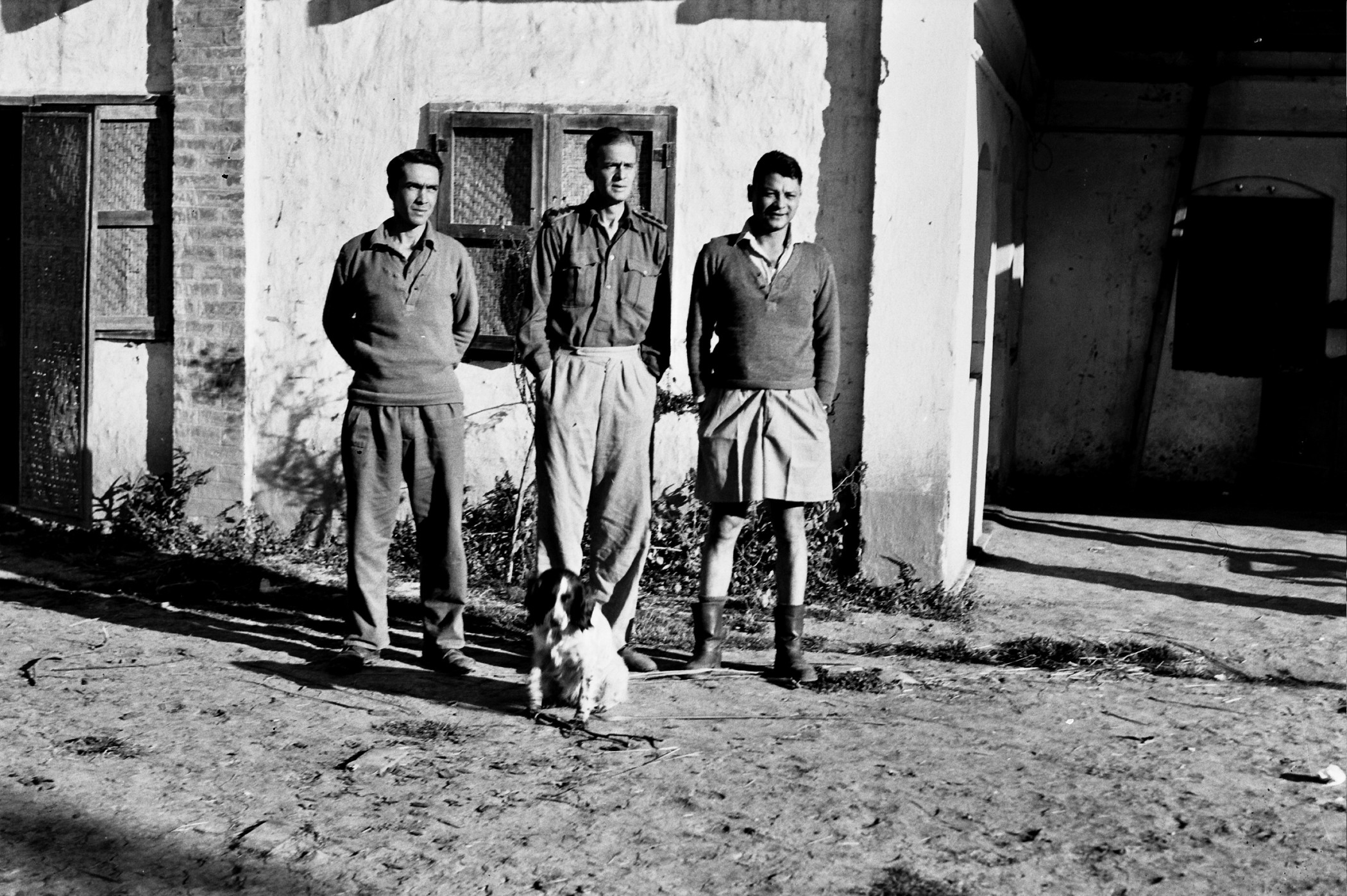 greyscale photo\' showing three men standing in a row in front of a white cottage-ish building, with a black and white spaniel sitting in front of them: on the left a swarthy man with his hands behind his back,wearing an army-style pullover and slacks; in the middle a tall white man with his hands in his pockets, wearing a military shirt and slacks; on the right a more Asian-looking man with his hands in his pockets, wearing a military shirt and shorts and Wellington boots