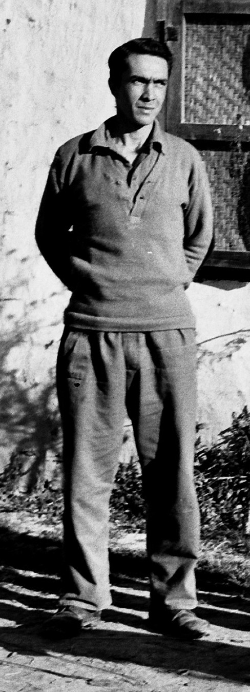 greyscale photo\' showing a swarthy man with his hands behind his back,wearing an army-style pullover and slacks