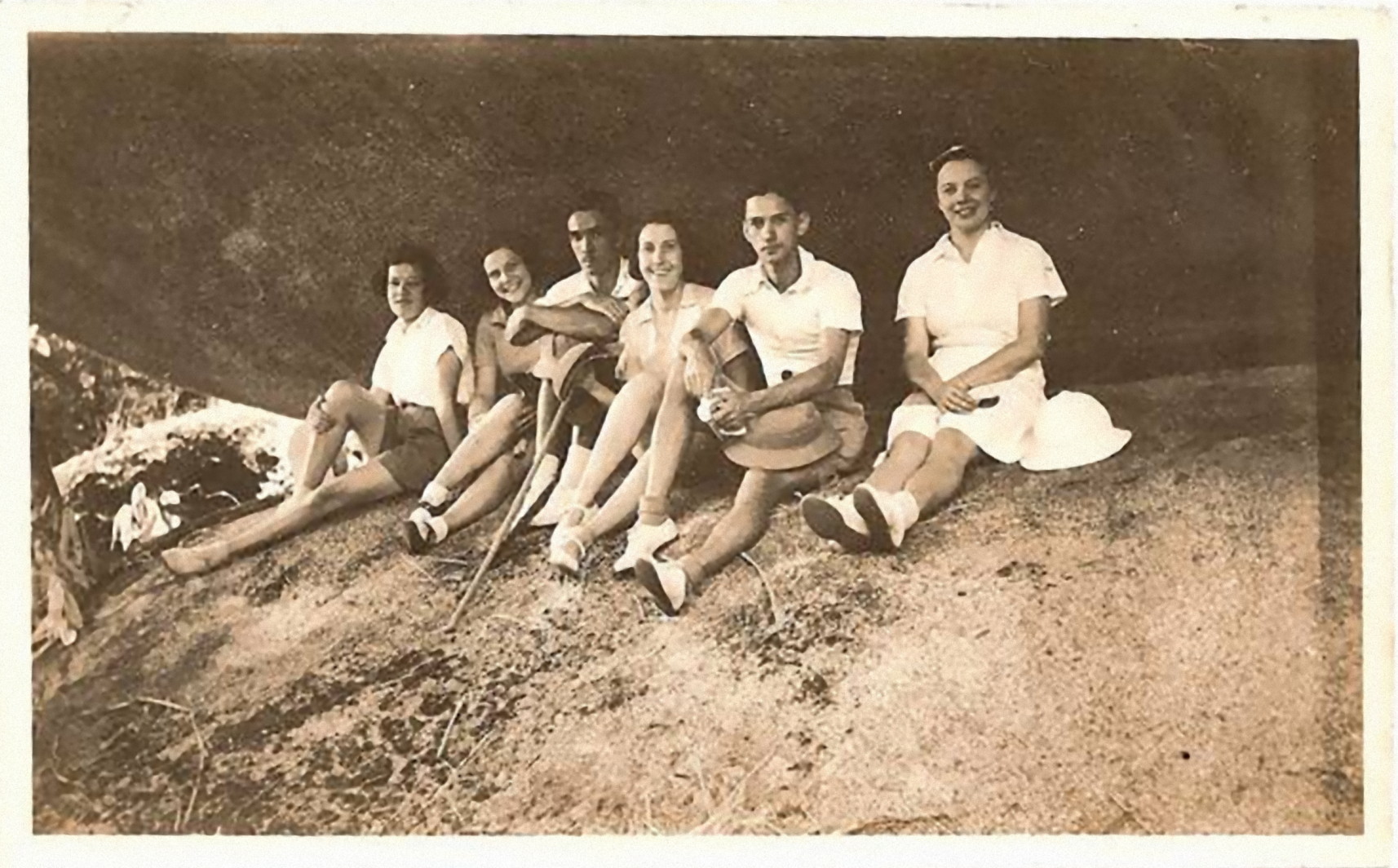 sepia photo\' showing six people all in about their twenties, dressed in lightweight summer clothes - from the left girl, girl, boy, girl, boy, girl - seated on an expanse of mossy rock with another huge curve of rock above them, rather as if they were sitting in the crack between two enormous bowling balls balanced one on top of the other
