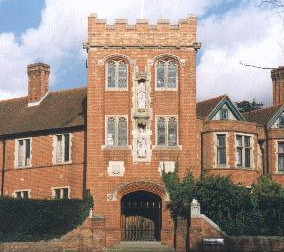 Victorian Gothic gateway in the form of a crenellated tower in ginger brick with an archway through it, with two storeys of pairs of Mediaeval-style windows above the arch and two stone statues of saints one above the other up the centre-front of the tower