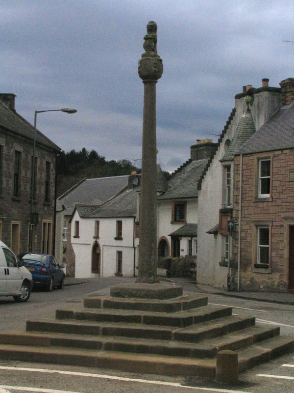 tall narrow stone pillar standing on top of a square pyramid of steps, with behind it a street of small old-fashioned buildings sloping down from the pillar