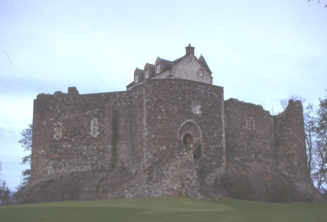 grey-brown curtain wall and gatehouse on top of a low mound, with the roof of a more house-like building visible over the top of the wall