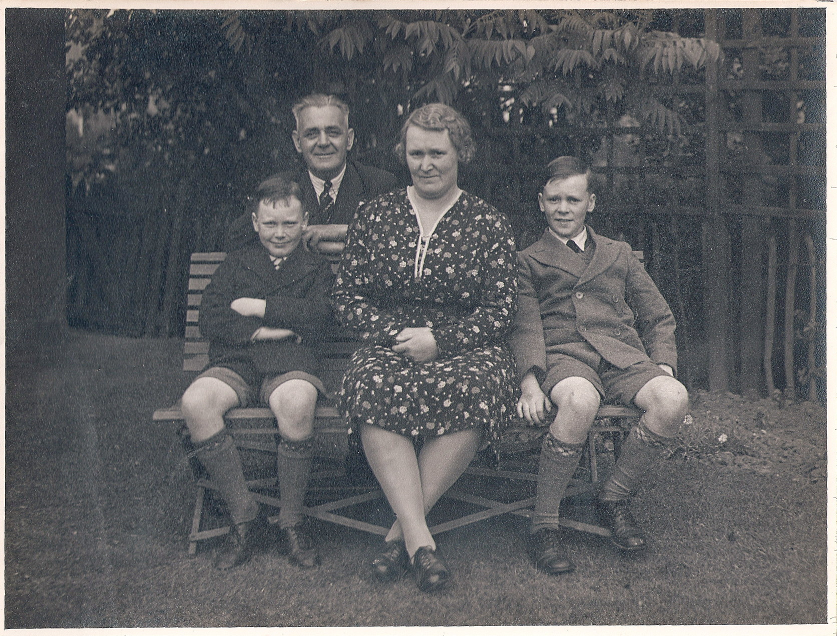 black and white photo\' showing a middle-aged woman with wavy brown hair and a dark, floral-print summer dress, seated on a garden bench and flanked by two boys aged about ten and wearing shool uniform, with a man with short, sticking-up grey hair leaning on the back of the bench
