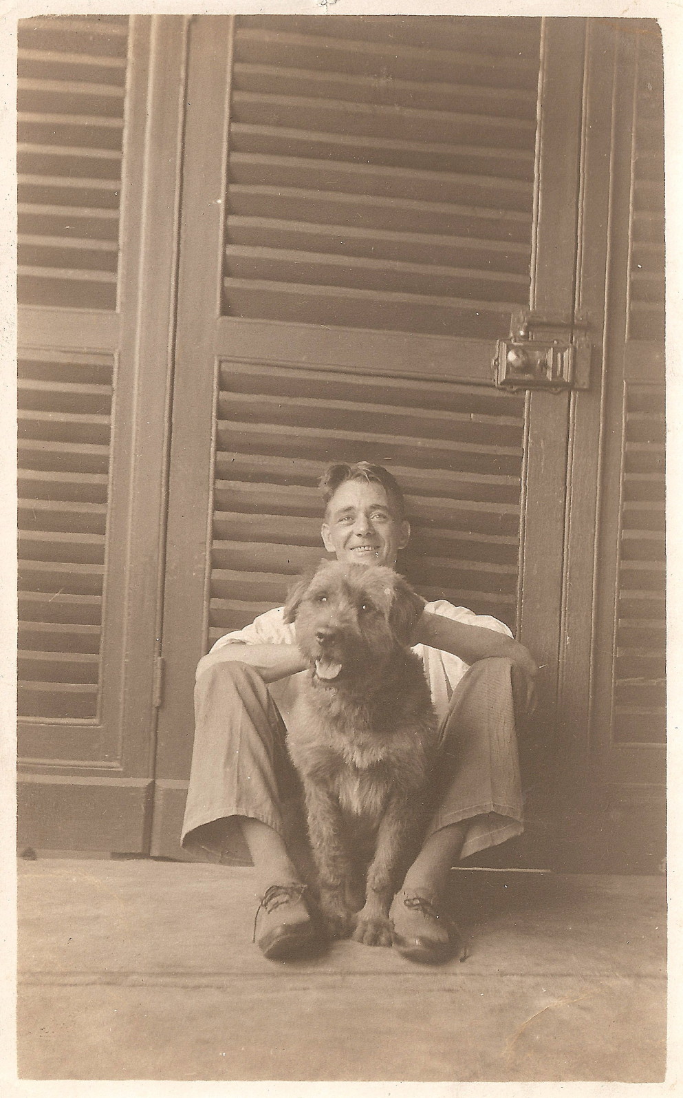 young man in a white shirt, pale floppy trousers and sandals, sitting on the ground in front of a slatted door, with a large shaggy dog between his knees