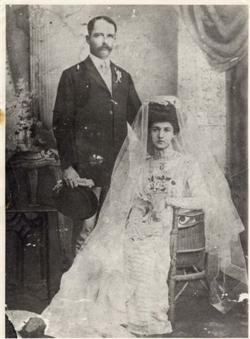 wedding photo\' showing an elegant woman in a slimline yet frothy, gauzy wedding dress, seated in a cane chair, with her husband in formal dress and a moustache standing at her right and holding a hat in his right hand
