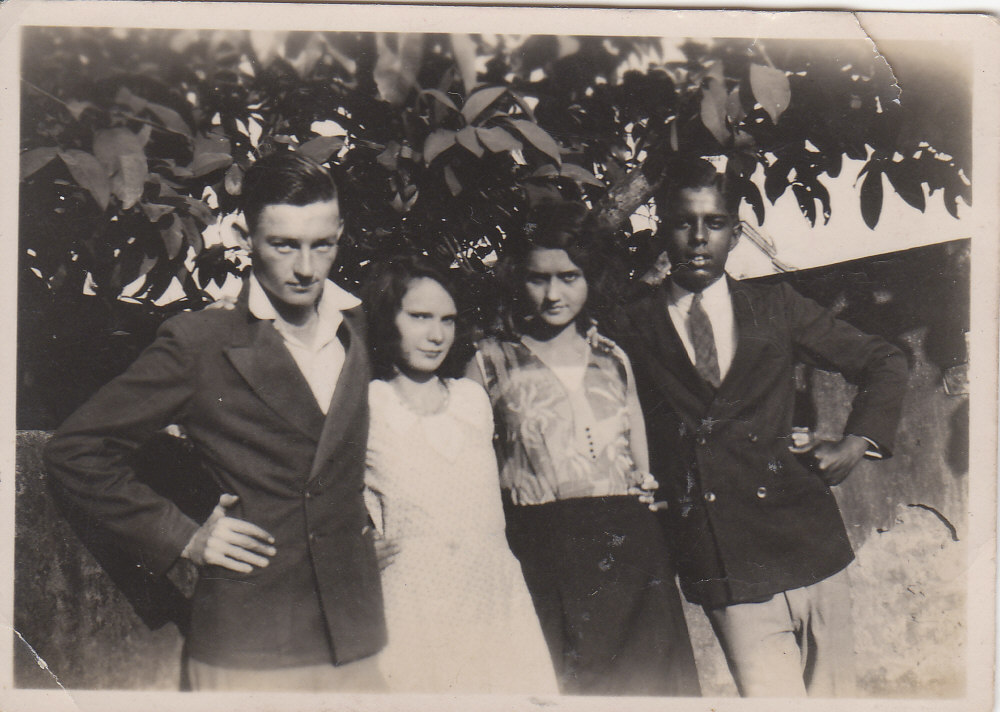 black and white photo\' of two boys, a white one on the left and a black one on the right, with two girls between them, all standing in front of bushes, both boys snappily-dressed and trying to look cool, and the girl to the right, next to the black boy, looking dark and exotic and sultry